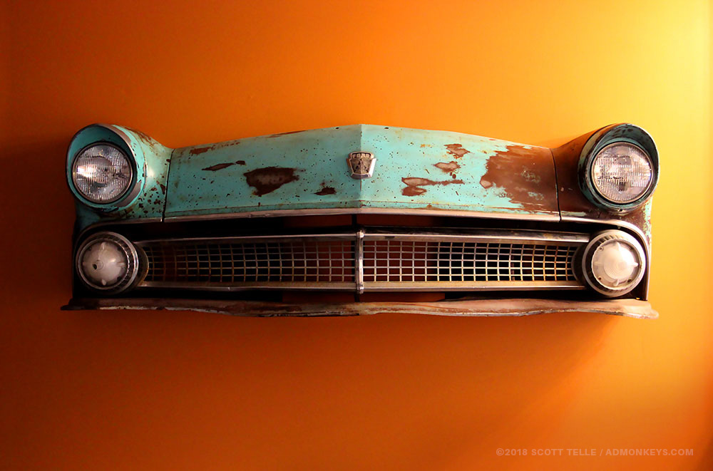 IMG_0769_ADM_Web_55FordCountrySedan_1000x661_200k+Tag The rusted front of a 1955 blue ford is hung up on an orange wall.
